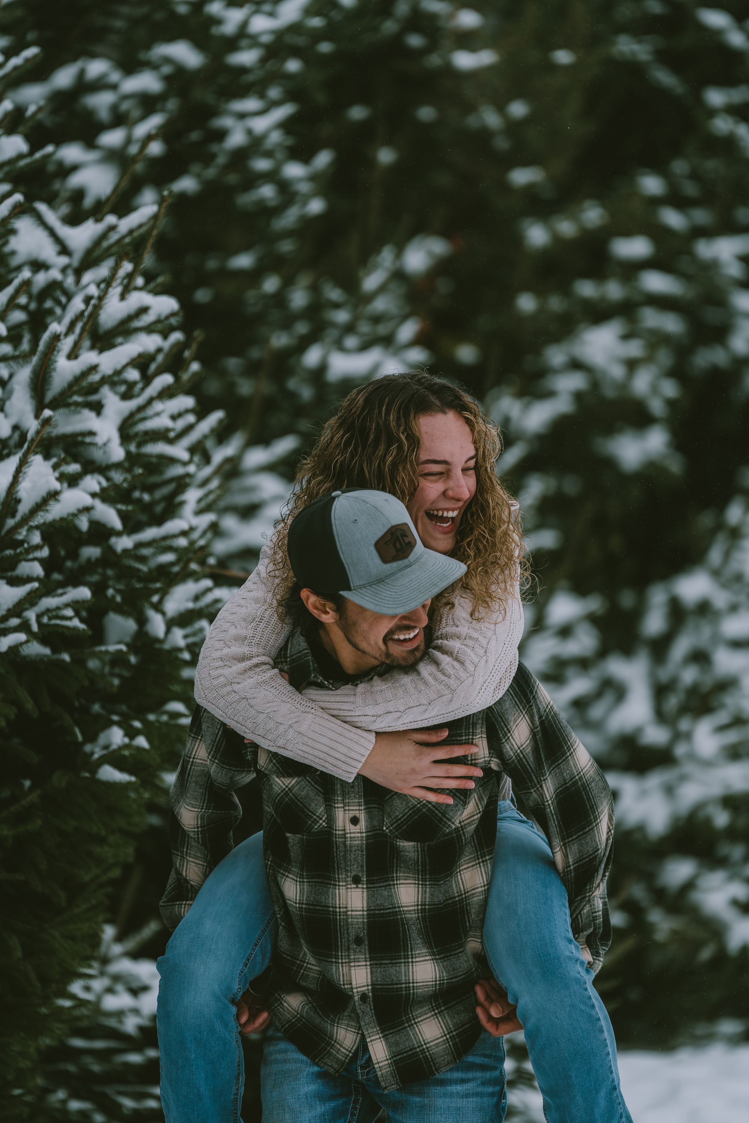 Joyful couple plays in snowy winter scene with person in plaid shirt giving piggyback ride through evergreen trees.