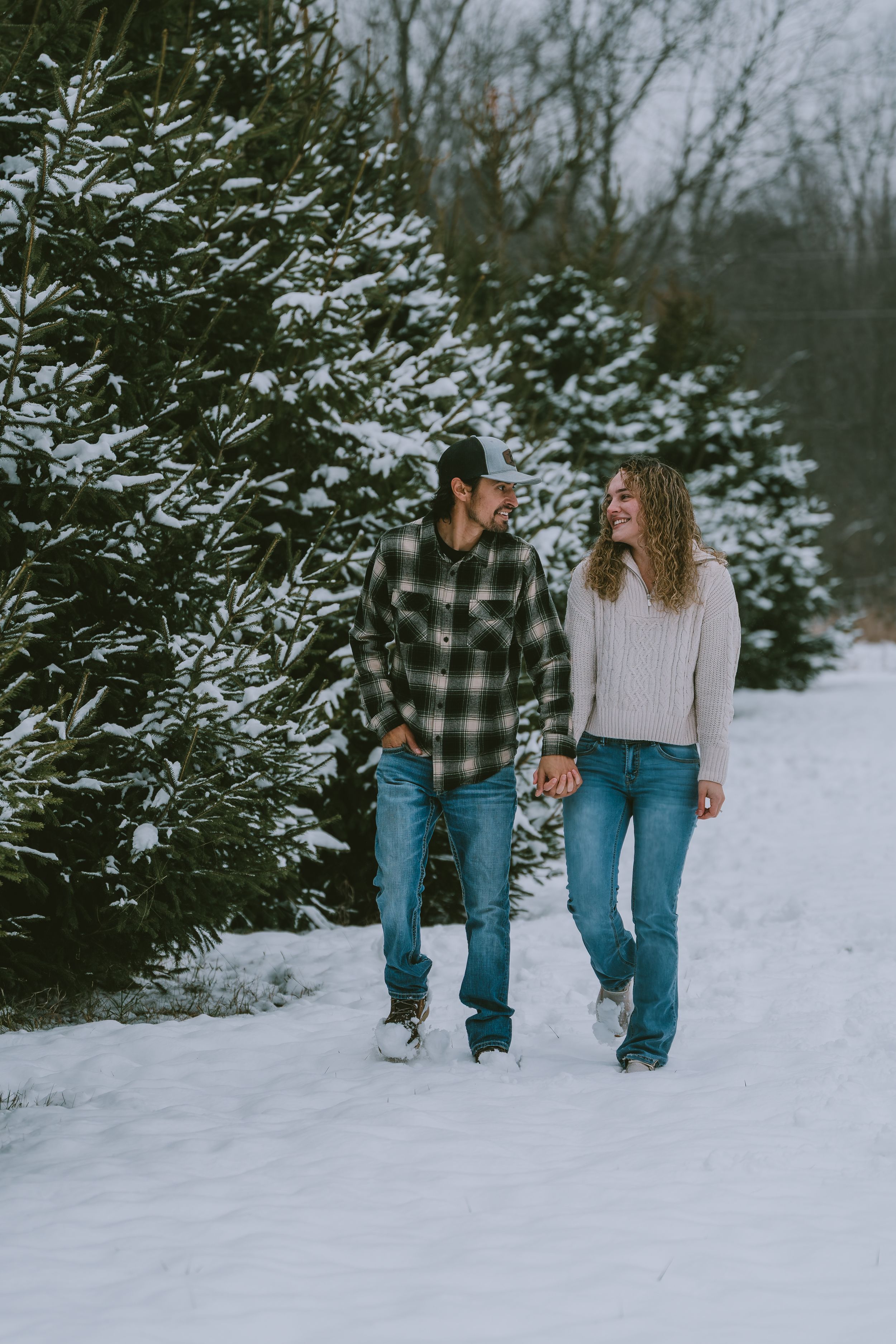 A couple walks hand in hand along a snowy path beside evergreen trees on a winter day.