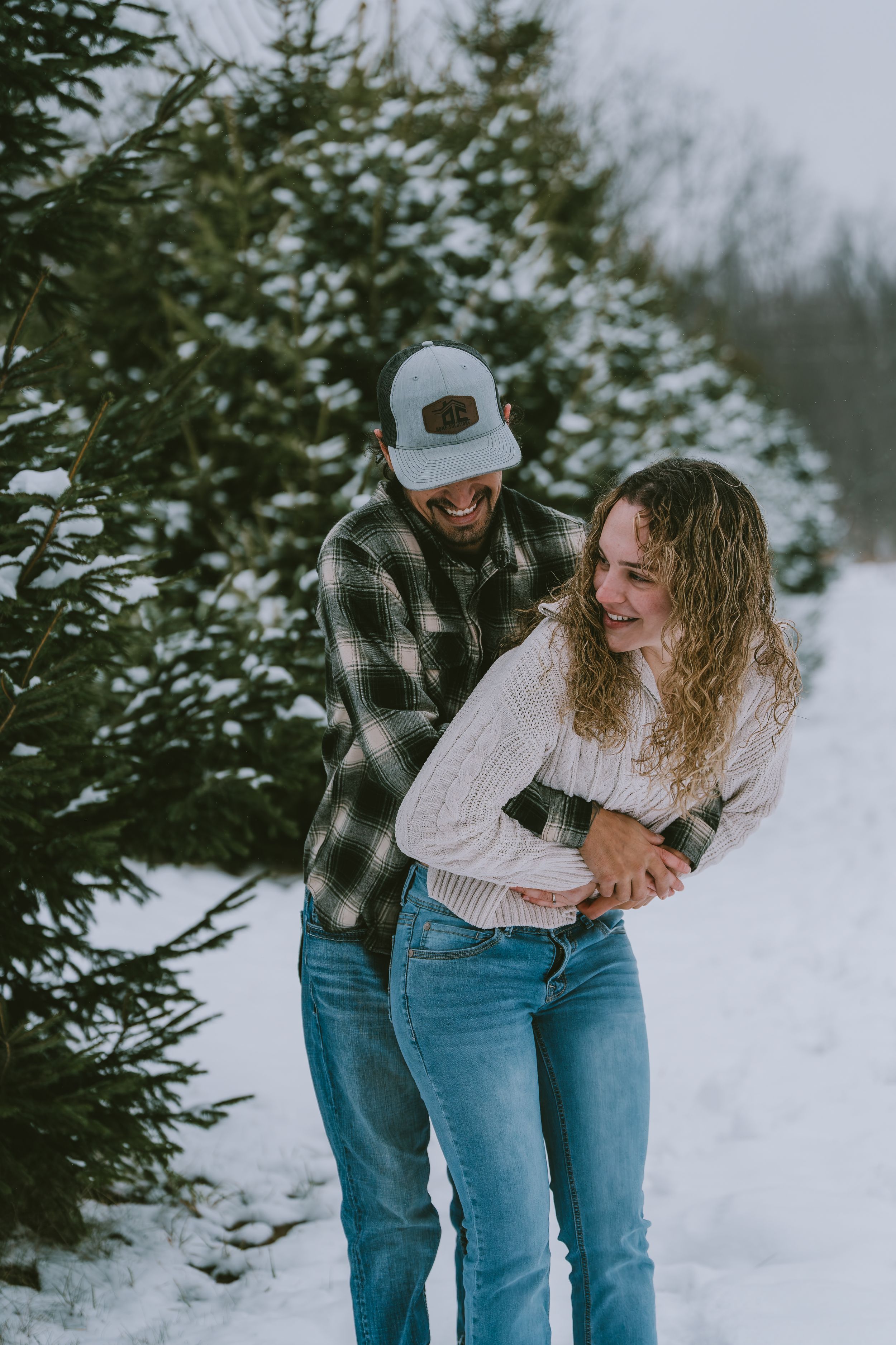 A couple plays and laughs together in the snow near evergreen trees on a winter day.