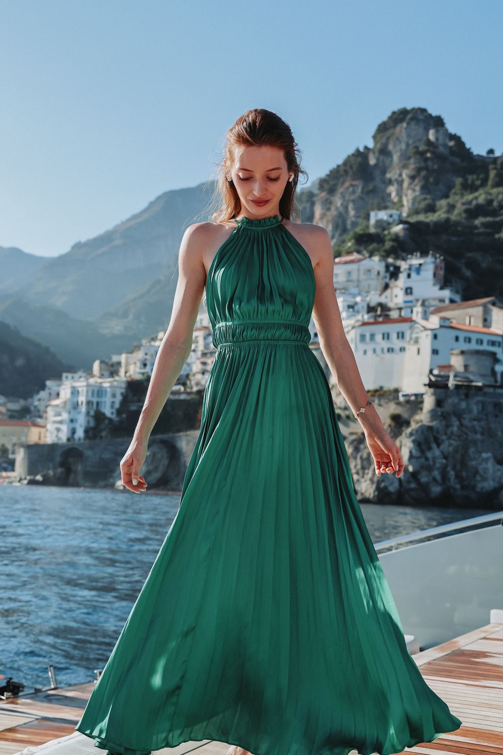 Woman in flowing green dress twirling on a boat deck with coastal Italian village backdrop.