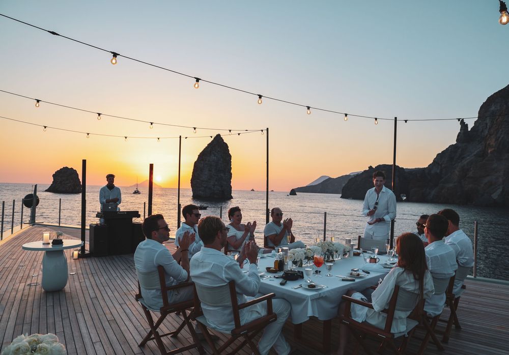 Sunset dinner party setup on a yacht deck with string lights and coastal scenery.