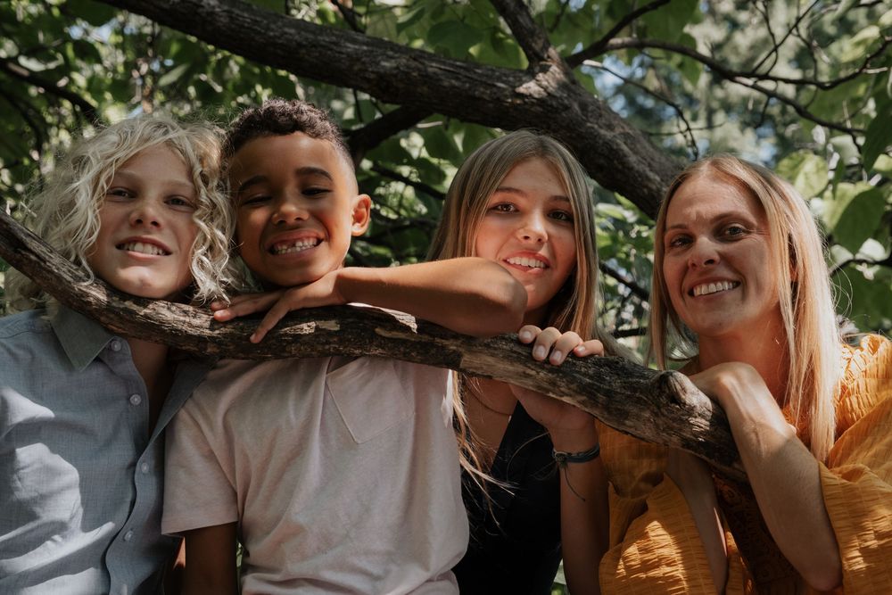 A smiling family embraces outdoors on a sunny day beneath green tree branches.