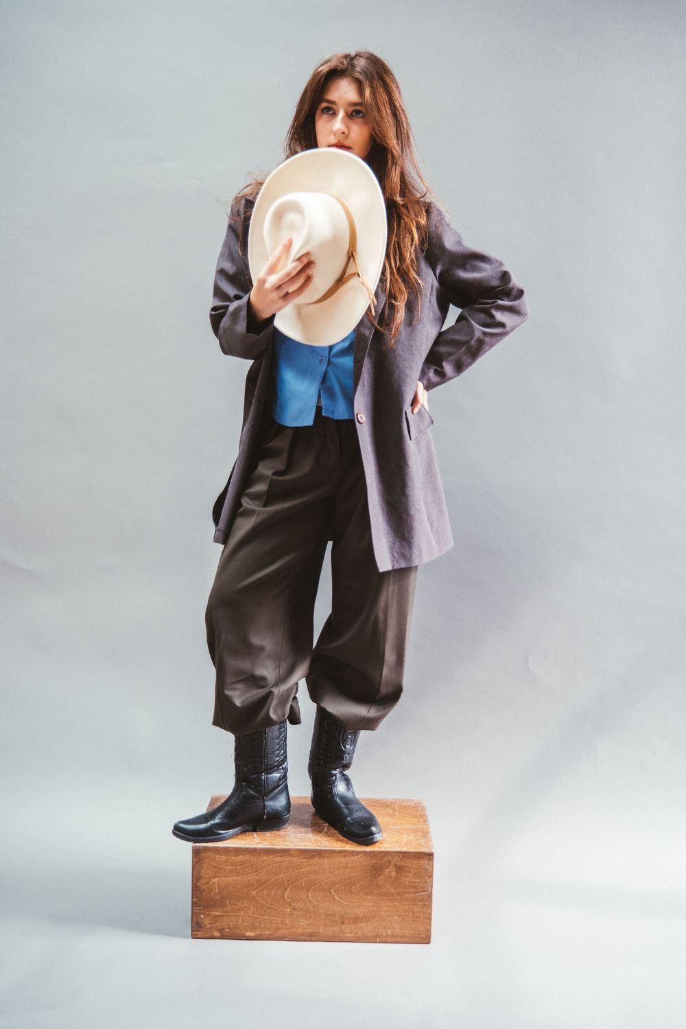 A stylish figure poses on a wooden block while wearing an oversized blazer, blue top and black shorts against a gray backdrop.