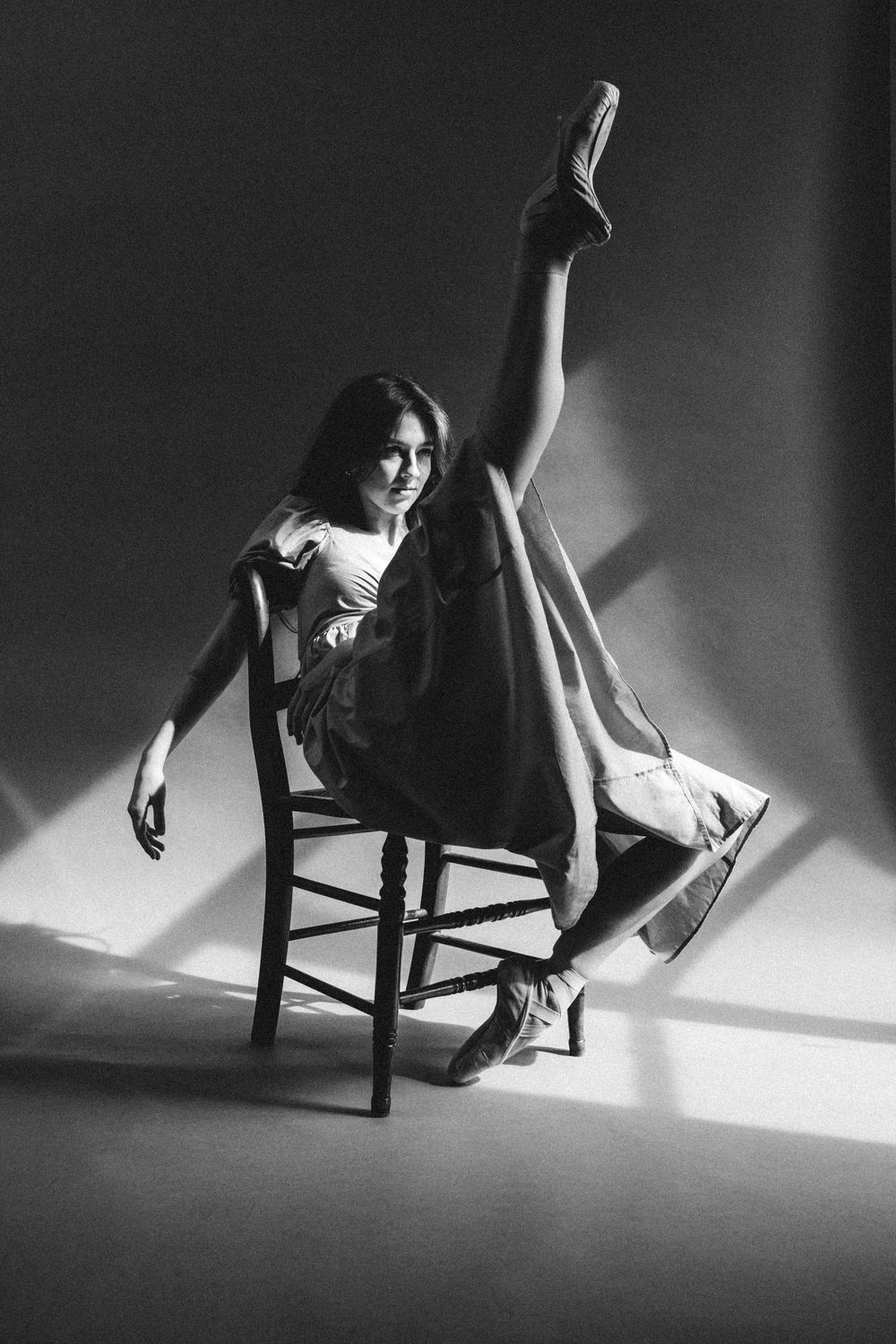 A dramatic black and white photograph shows a dancer posing gracefully on a wooden chair with one leg extended upward.