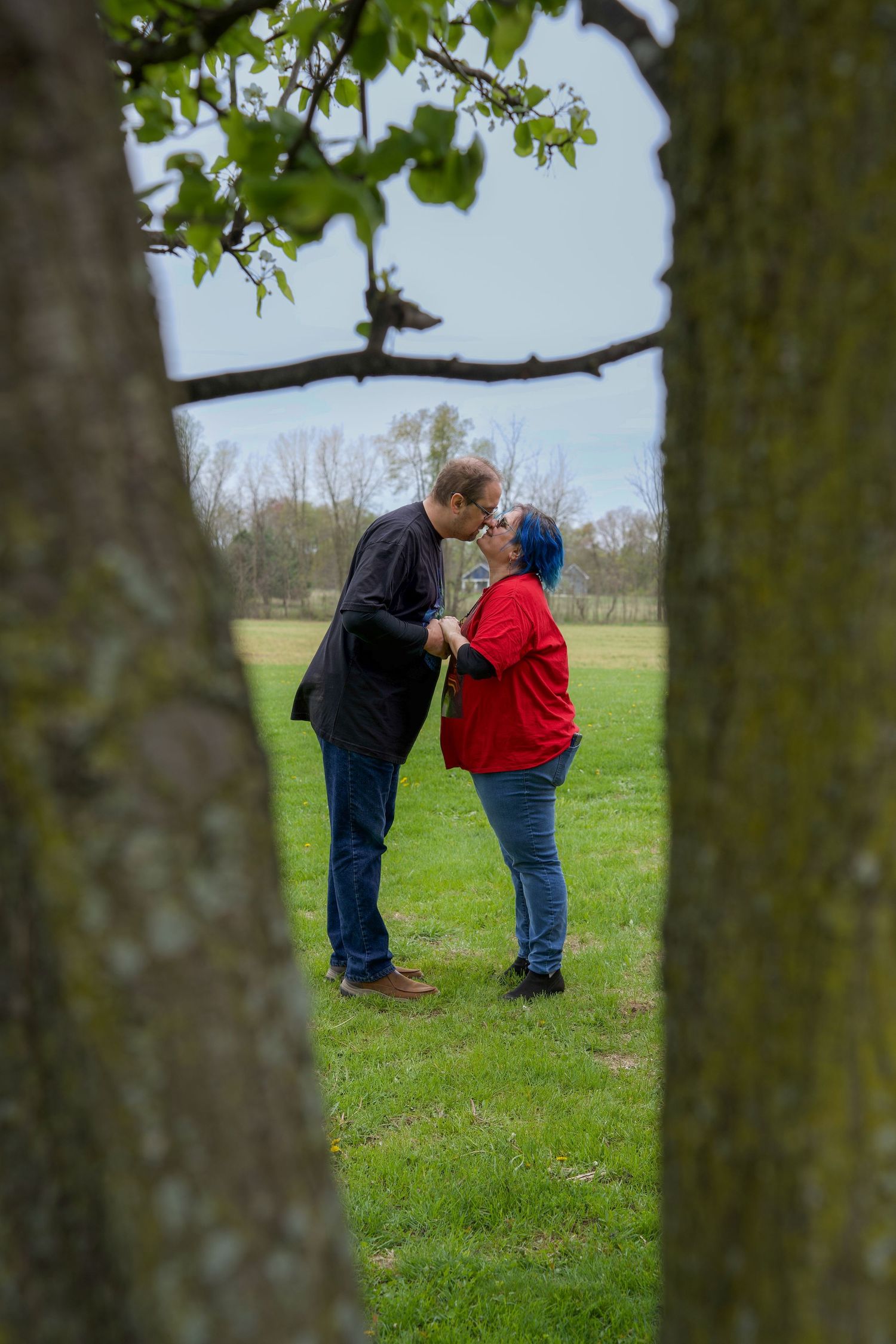 A couple shares an intimate moment while standing together in a grassy field, viewed through tree trunks in the foreground.