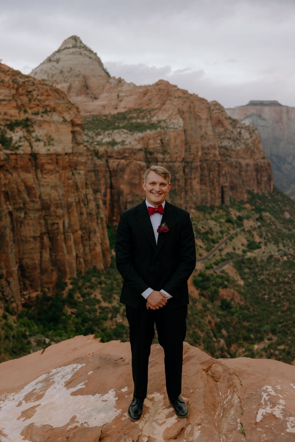 A person in a black formal suit standing on red rocks with dramatic canyon cliffs in the background at sunset.