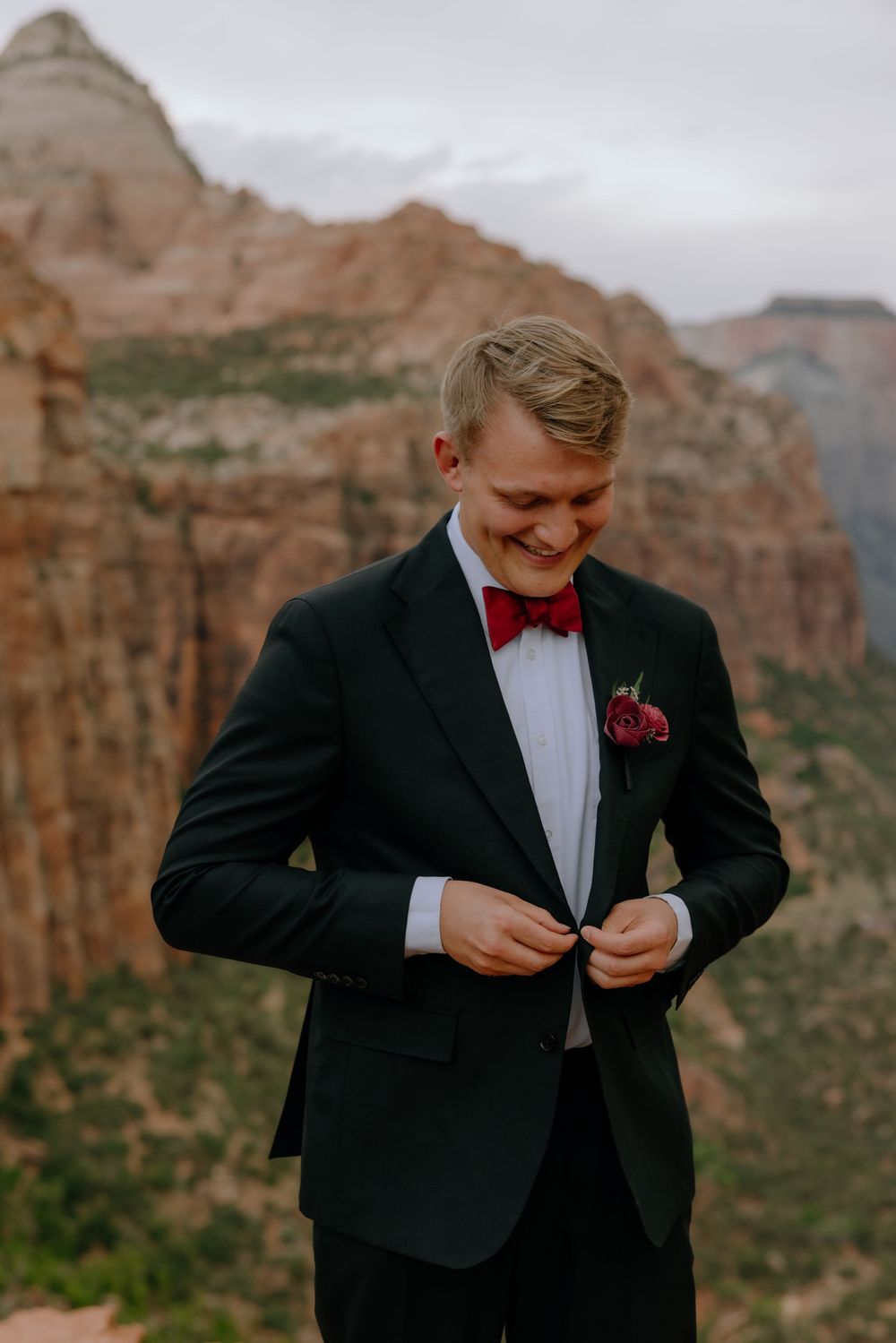 Groom in black tuxedo and red bow tie smiles while adjusting outfit against dramatic red rock canyon backdrop.