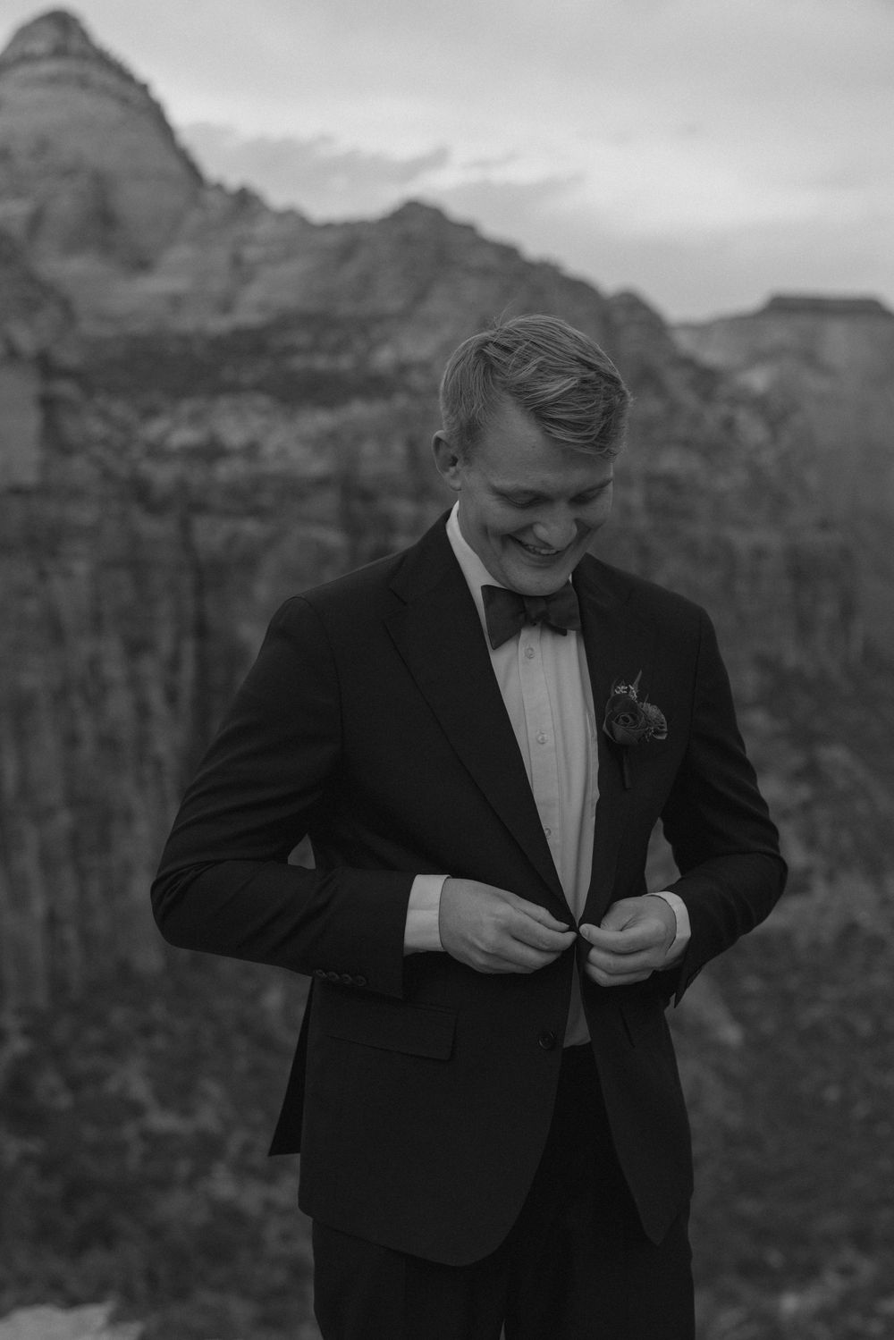 A person in a black tuxedo and bowtie smiles while adjusting their jacket against a mountainous landscape background.