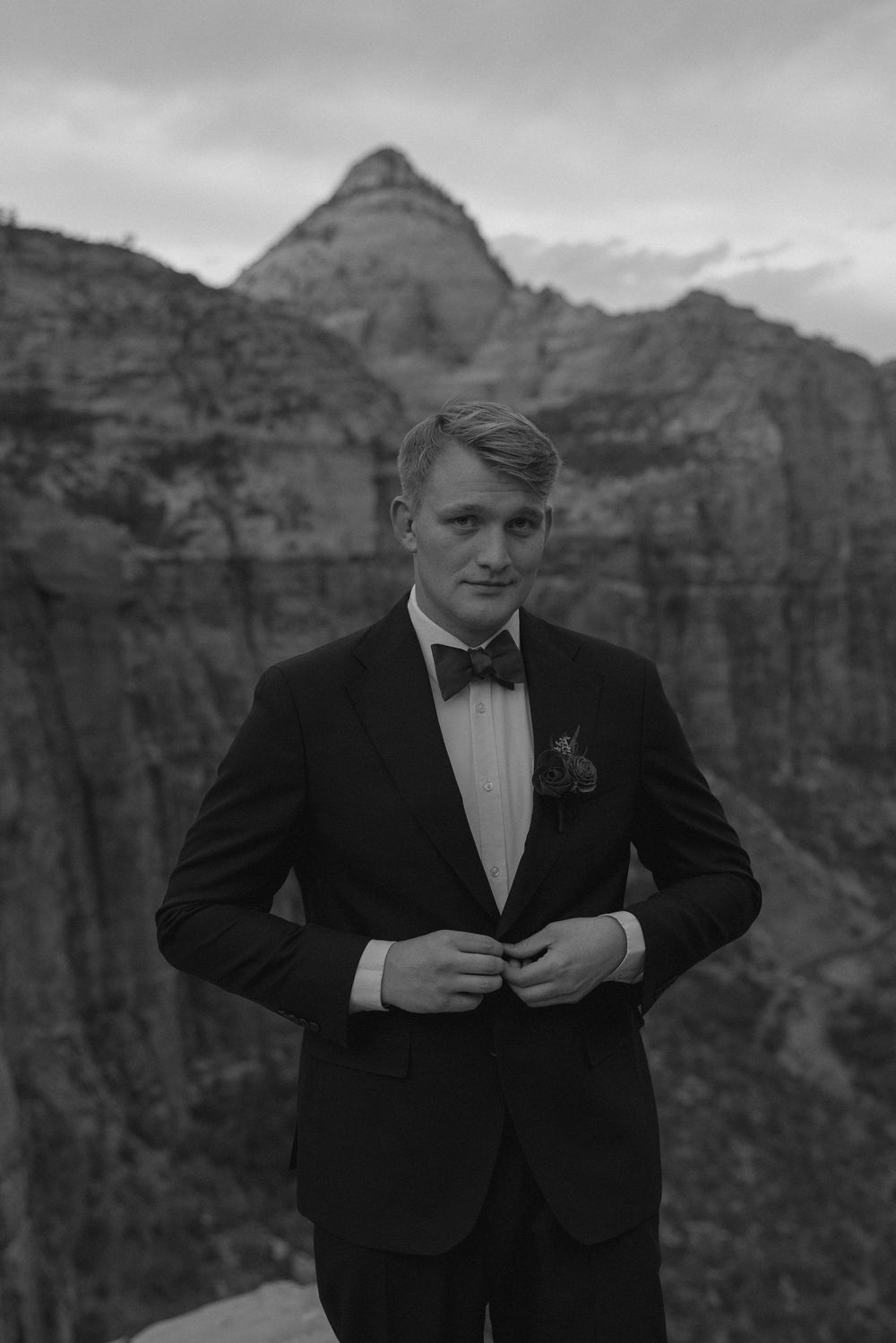 Classic black and white portrait of a groom in a tuxedo standing against dramatic canyon cliffs at sunset.