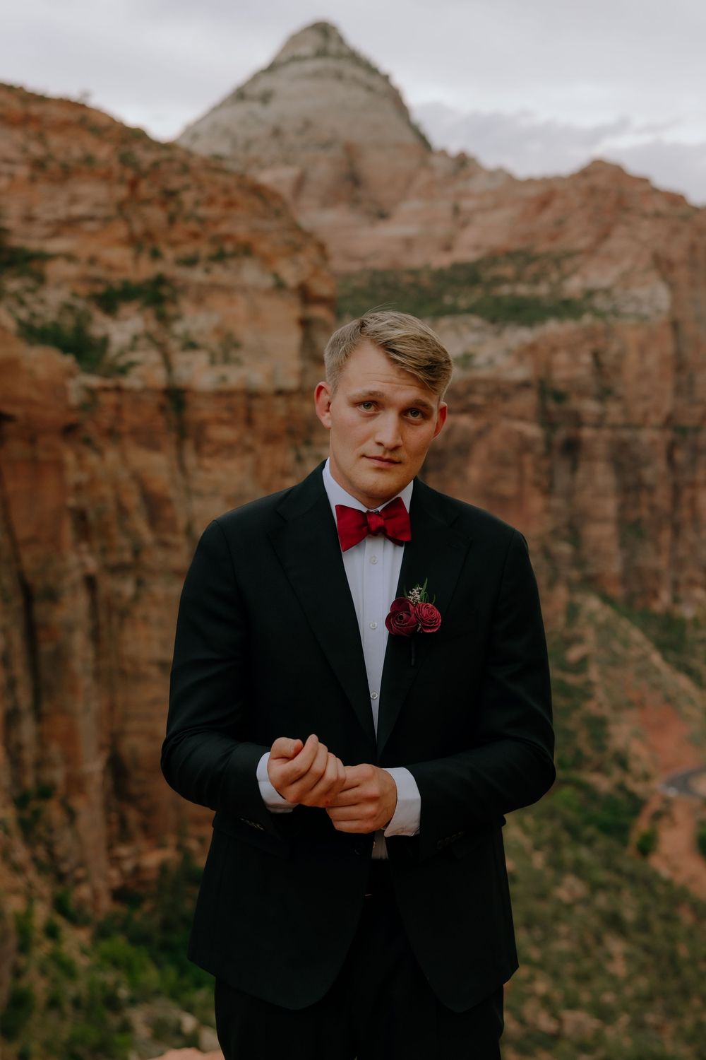 Formal portrait of a person in a black tuxedo and red bowtie standing against dramatic red rock cliffs at sunset.