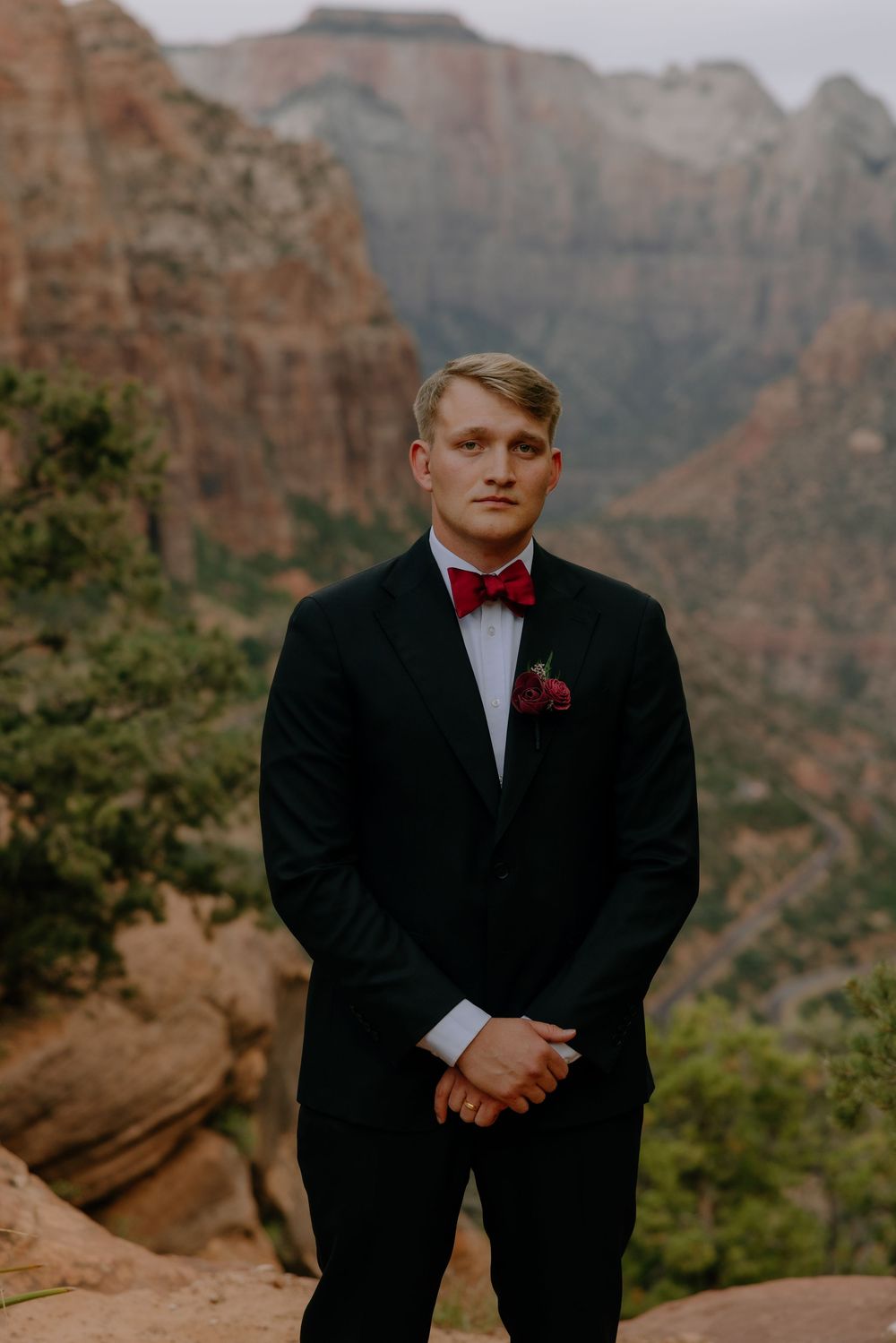 Well-dressed groom in black tuxedo and red bow tie poses against dramatic red rock cliffs in Zion National Park.