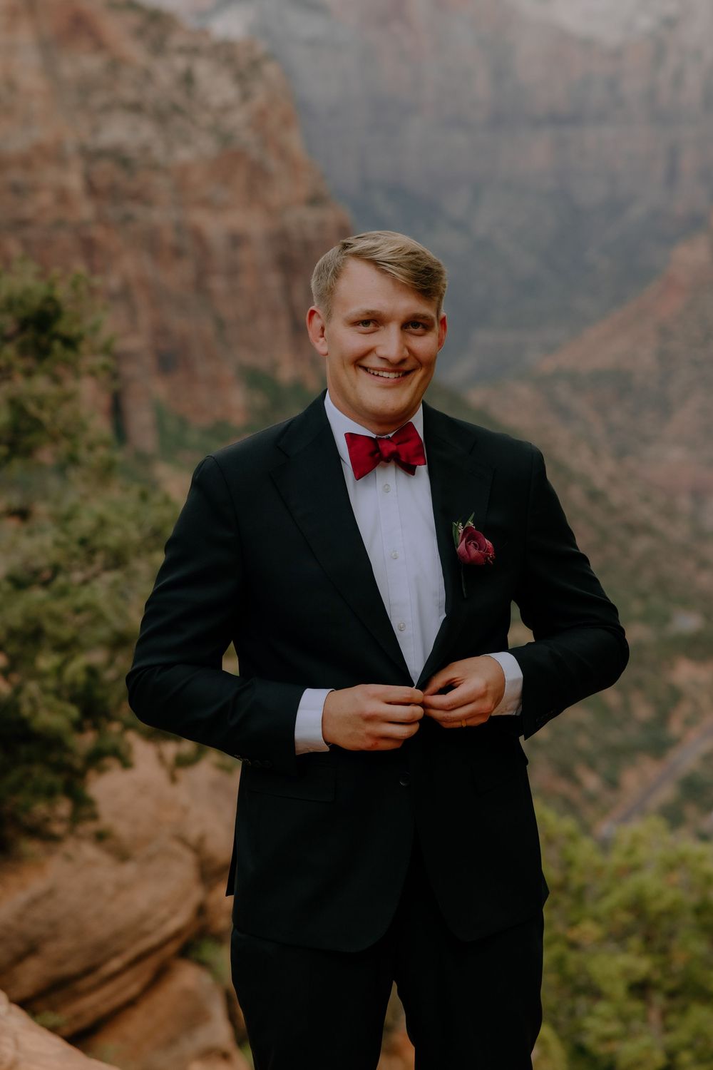 A person dressed in a black tuxedo with a red bow tie smiles while standing against stunning canyon scenery.