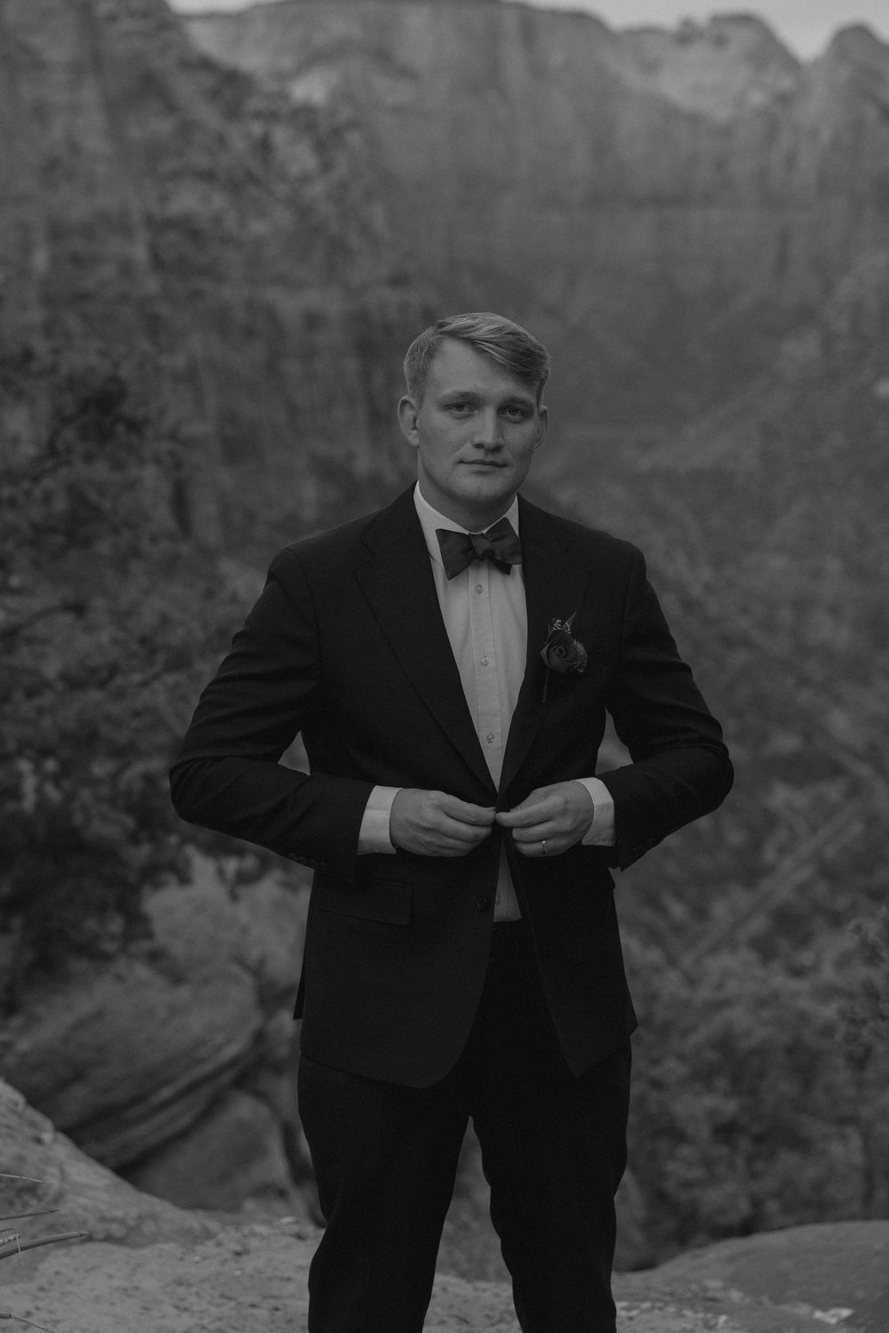 A groom in formal black tuxedo adjusts his bowtie while standing on a cliff overlook in Zion National Park.