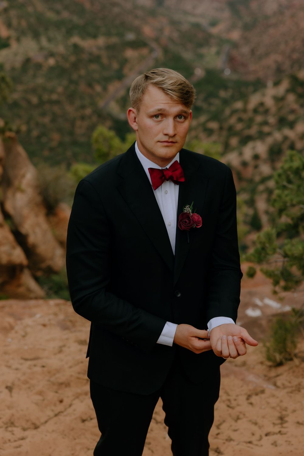 A groom in a black tuxedo and red bow tie poses against a scenic mountainous backdrop in warm lighting.