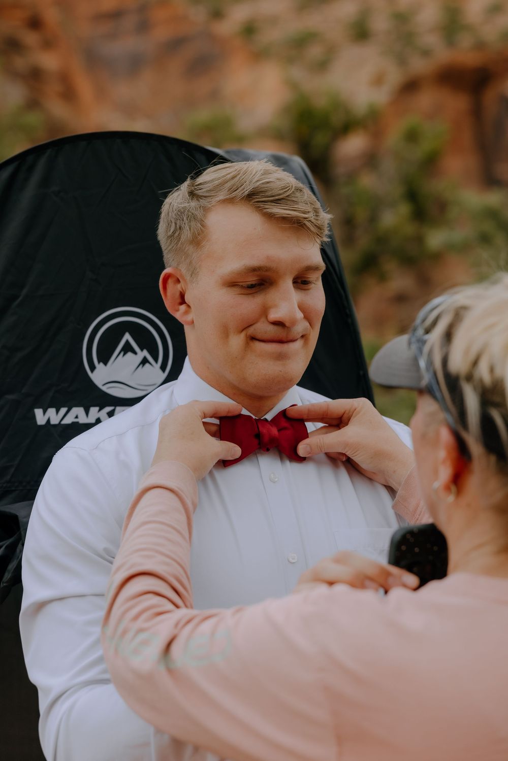Someone helps adjust a red bowtie on a groom wearing a white dress shirt during what appears to be a wedding preparation.