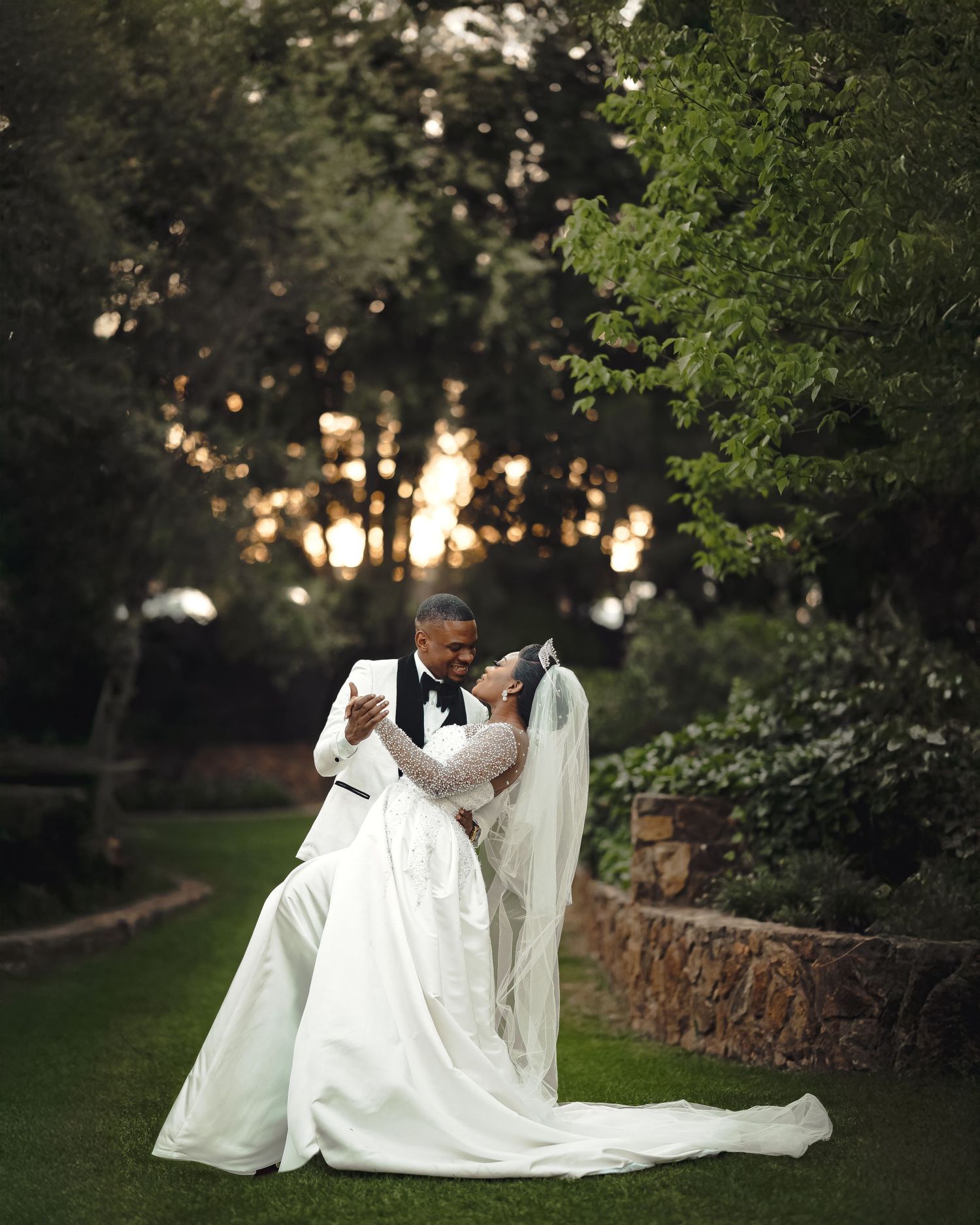 A romantic wedding couple embraces in a beautiful garden setting with soft sunset lighting filtering through the trees.
