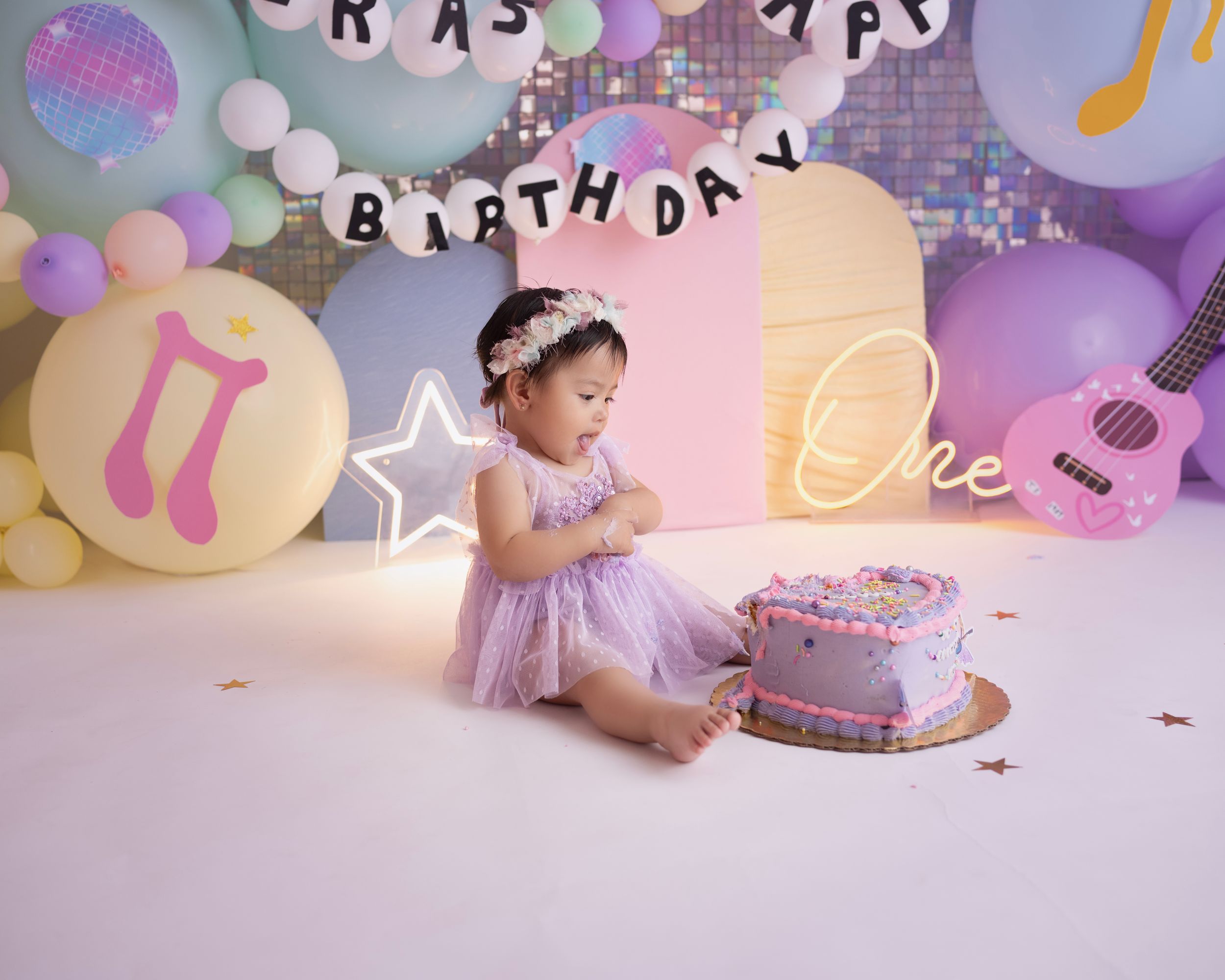 First birthday photo with purple cake and pastel balloon decorations.