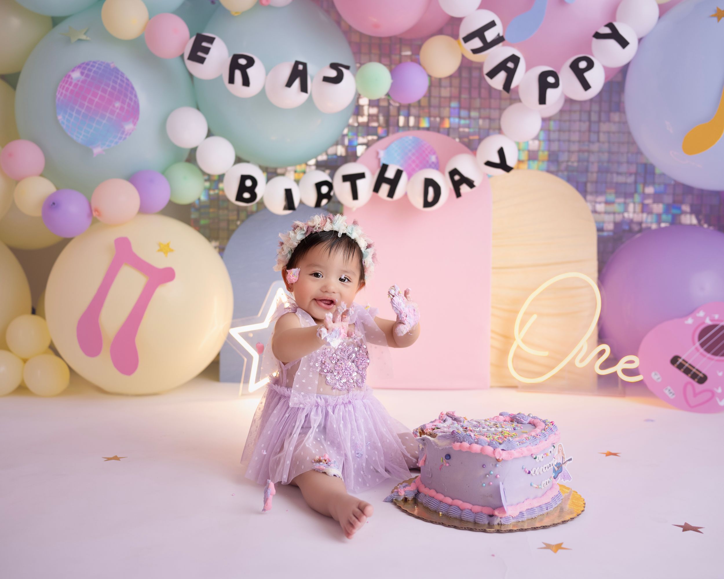 First birthday celebration setup with pastel balloons, 'Happy Birthday' banner and purple cake against a pink backdrop.