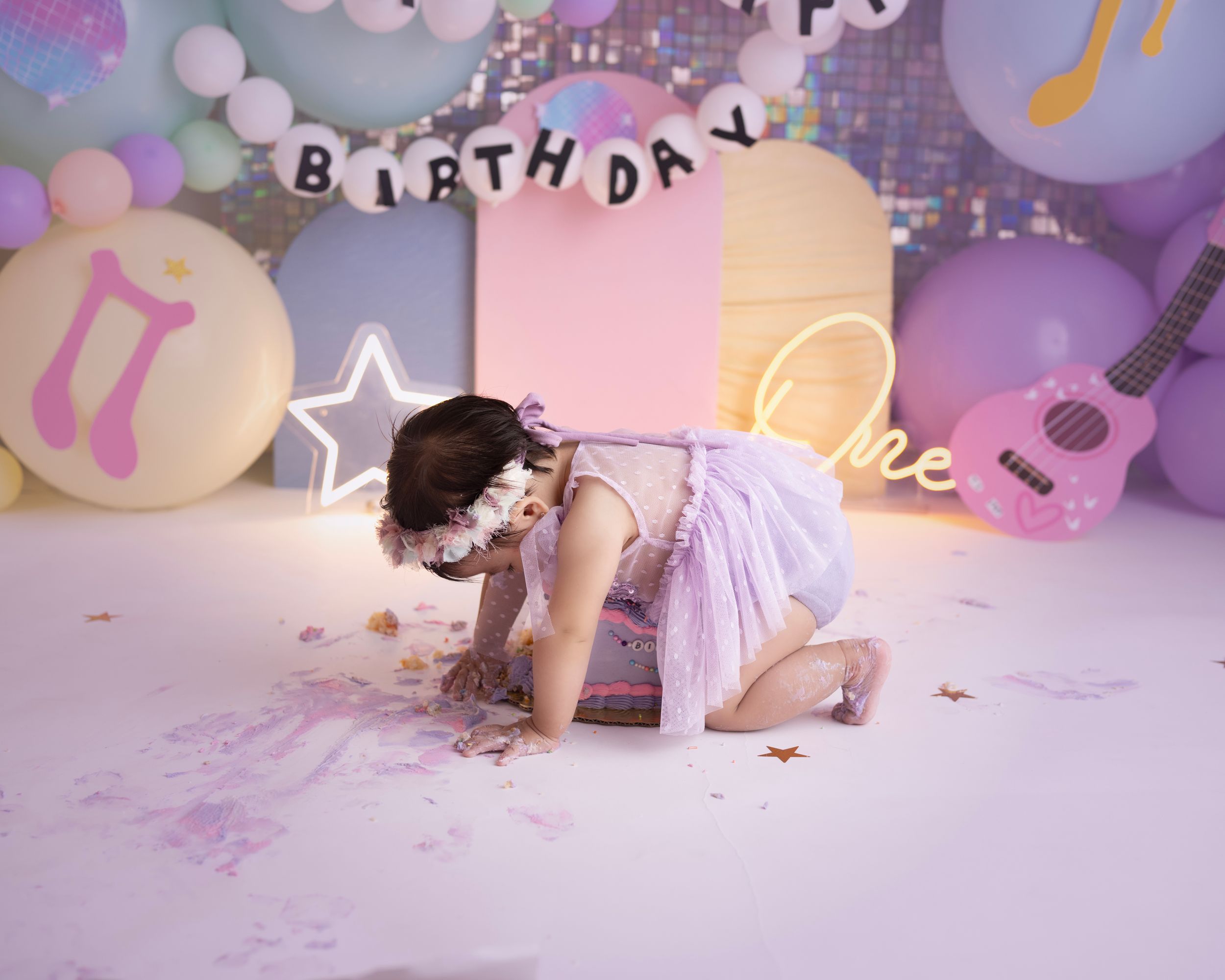 A festive first birthday cake smash scene with purple and pink balloons, musical decorations and a star light.