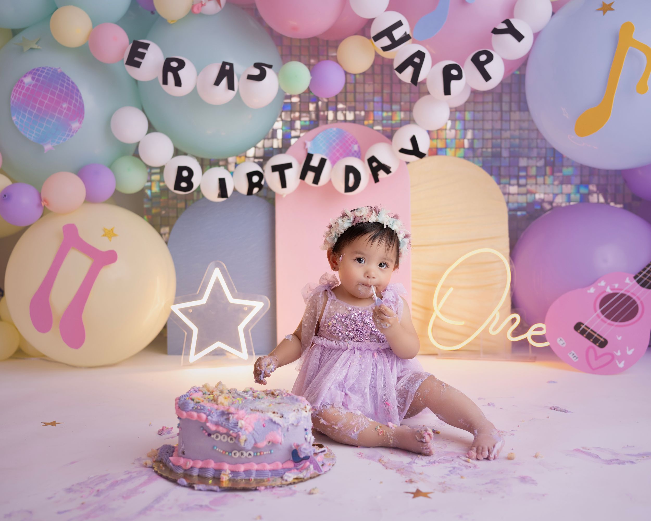 A festive pink and purple first birthday party setup with balloons, musical decorations, a cake and "Happy Birthday" banner.
