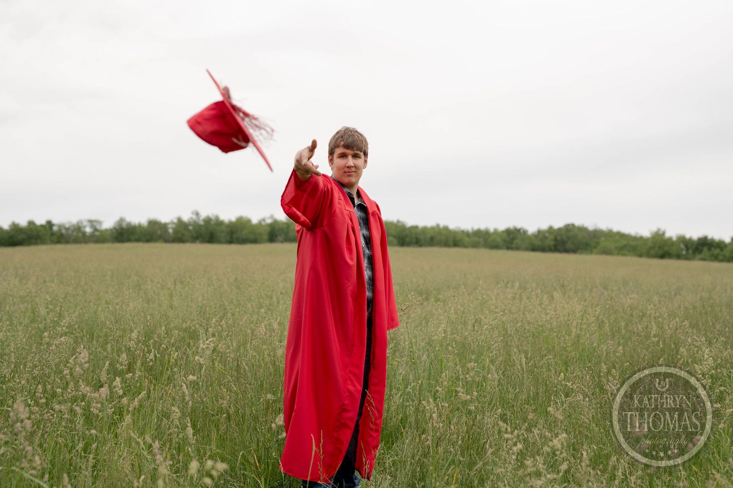 Graduate in bright red cap and gown stands triumphantly in a green meadow field holding their graduation cap.