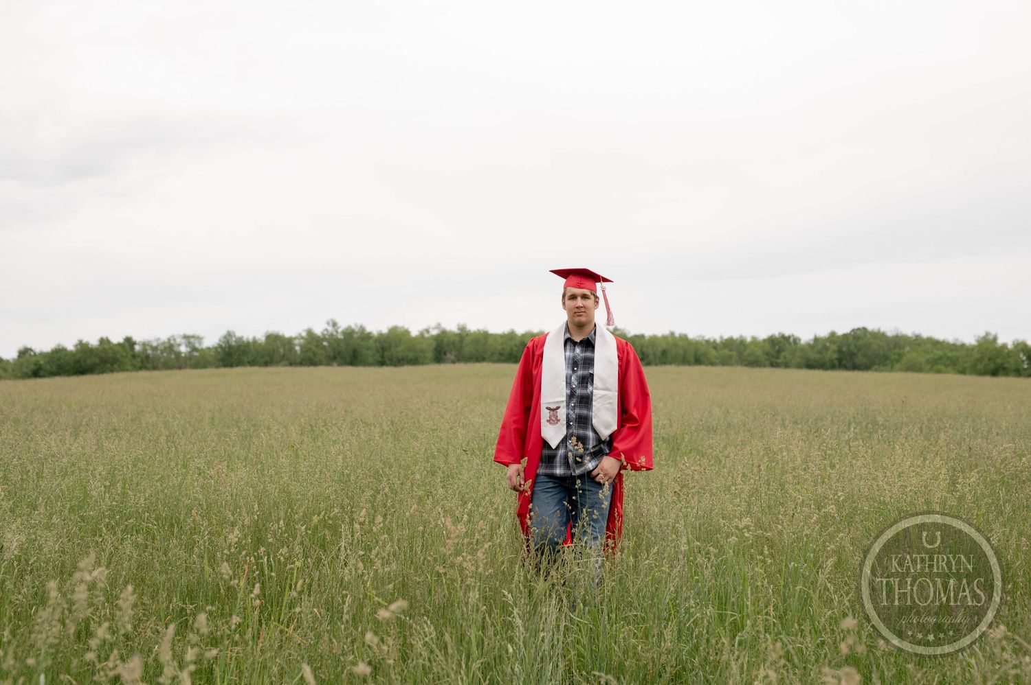 Graduate in red cap and gown stands in an open green field on a cloudy spring day.