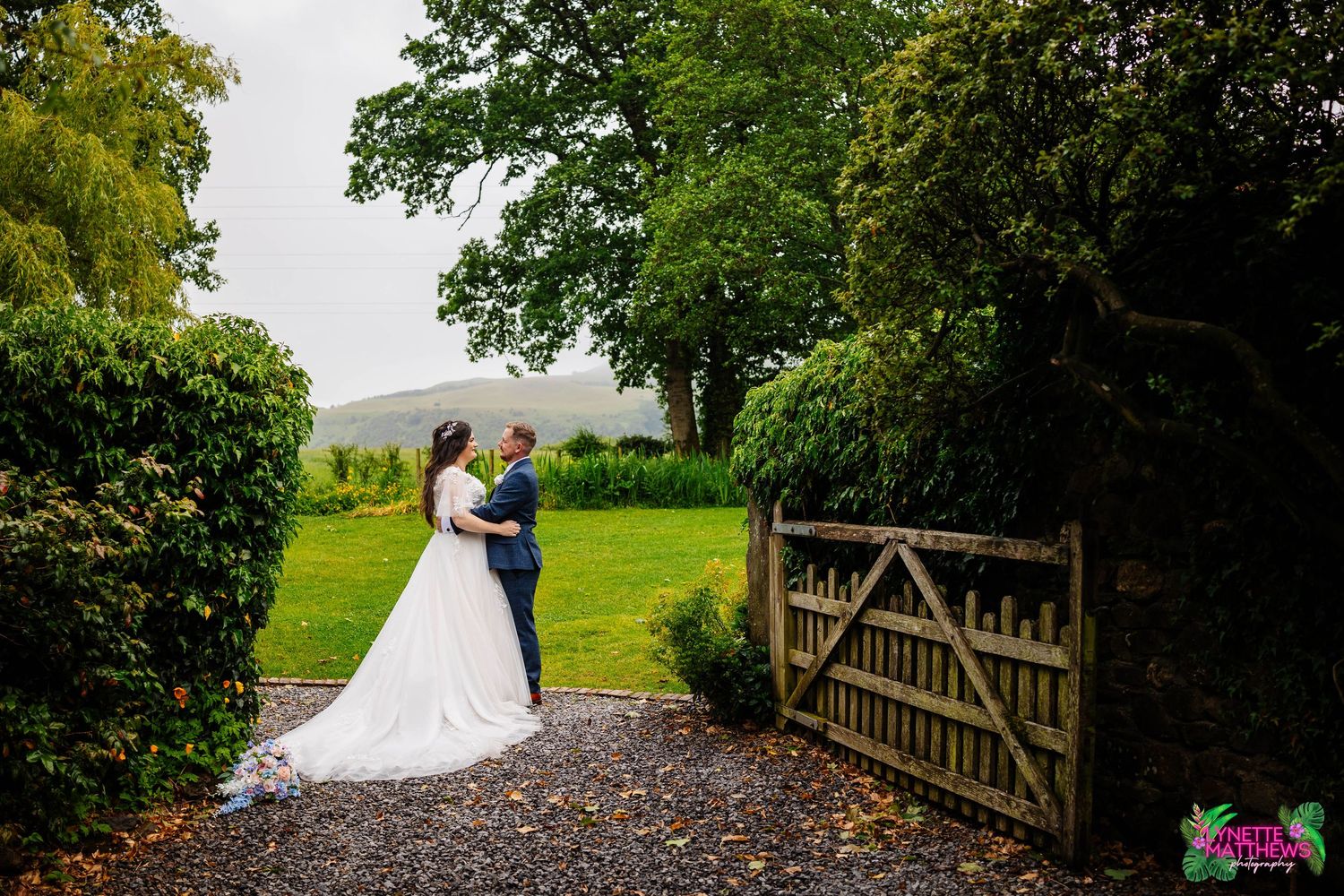 A romantic countryside wedding scene with a wooden gate, vibrant green trees & hills on a rainy wedding day at Low Hall by Lynette Matthews
