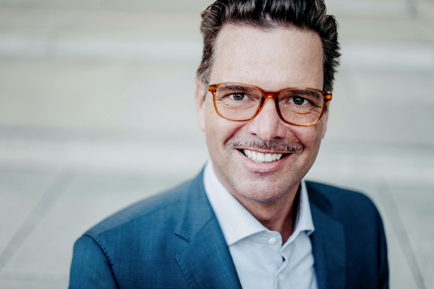 Eine lächelnde Person mit Brille und Schnurrbart trägt einen blauen Anzug und steht vor einem unscharfen Hintergrund.