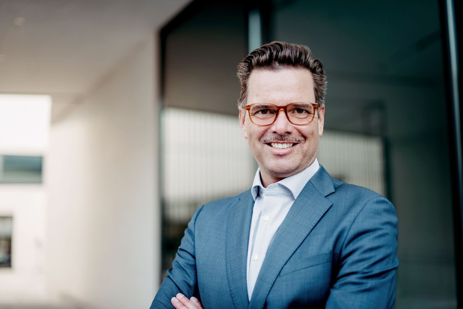 Businessfoto eines Mannes mit Blauem Anzug, weissem Hemd und Brille. Er lächelt in die Kamera. Im Hintergrund ist eine Wand mit Glastüren.