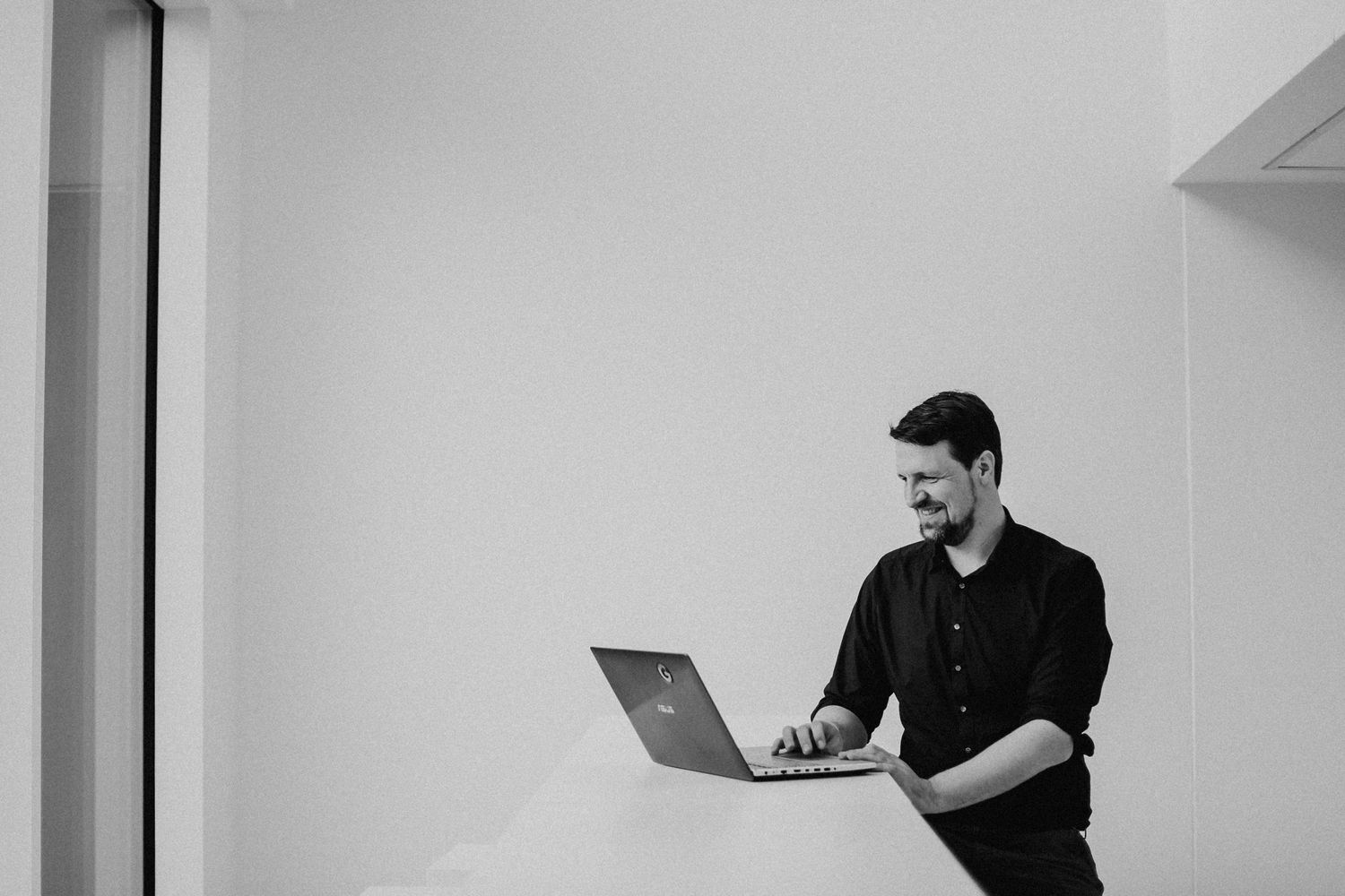 Ein Mann steht an einem Tisch und arbeitet an einem Laptop. Der Raum ist minimalistisch gestaltet und in Schwarz-Weiß gehalten.