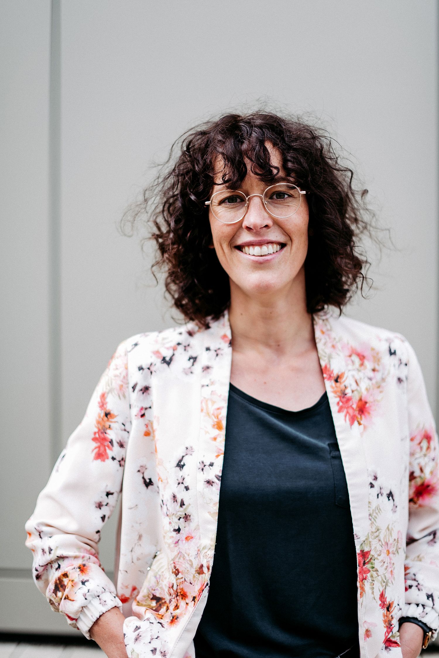 Portrait einer Frau mit braunen lockigen Haaren. Sie trägt eine Brille und eine Jacke mit floralem Muster.