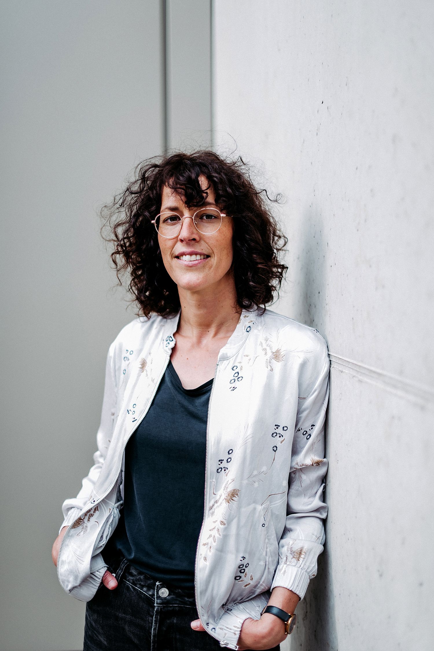 Frau mit lockigem Haar und Brille lehnt lächelnd an einer Betonwand. Sie trägt eine helle Jacke und ein dunkles Shirt.