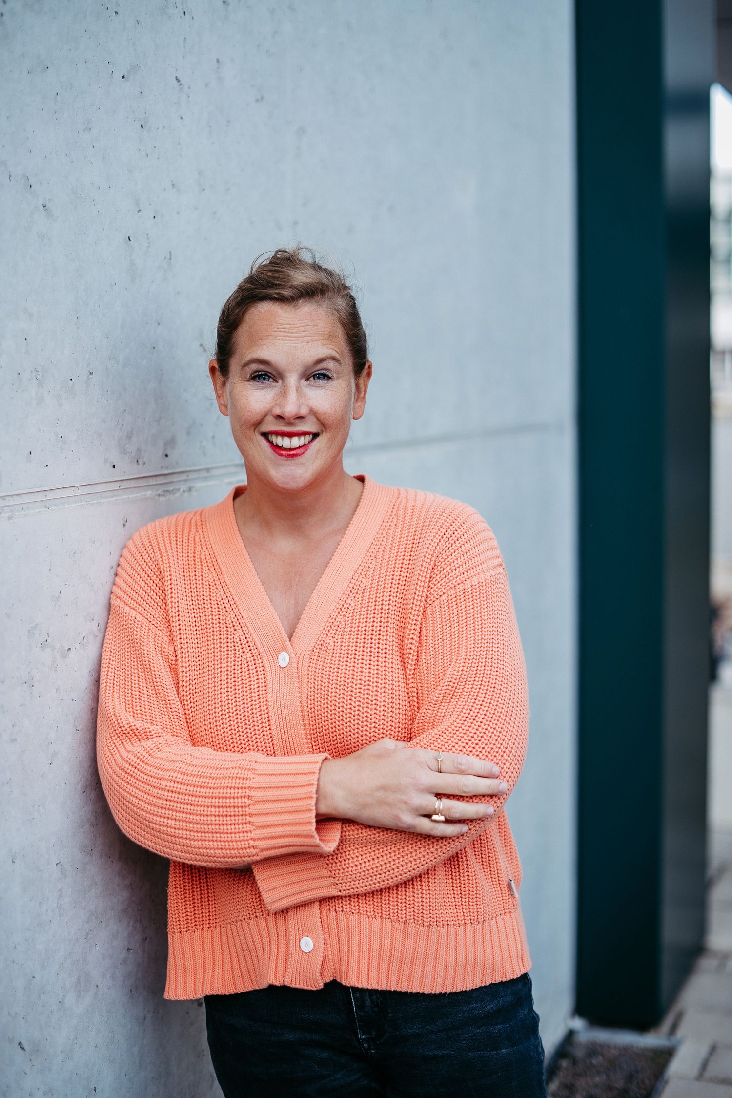 Frau lächelt in die Kamera. Sie trägt einen orangefarbenen Strickpullover und steht vor einer Betonwand.