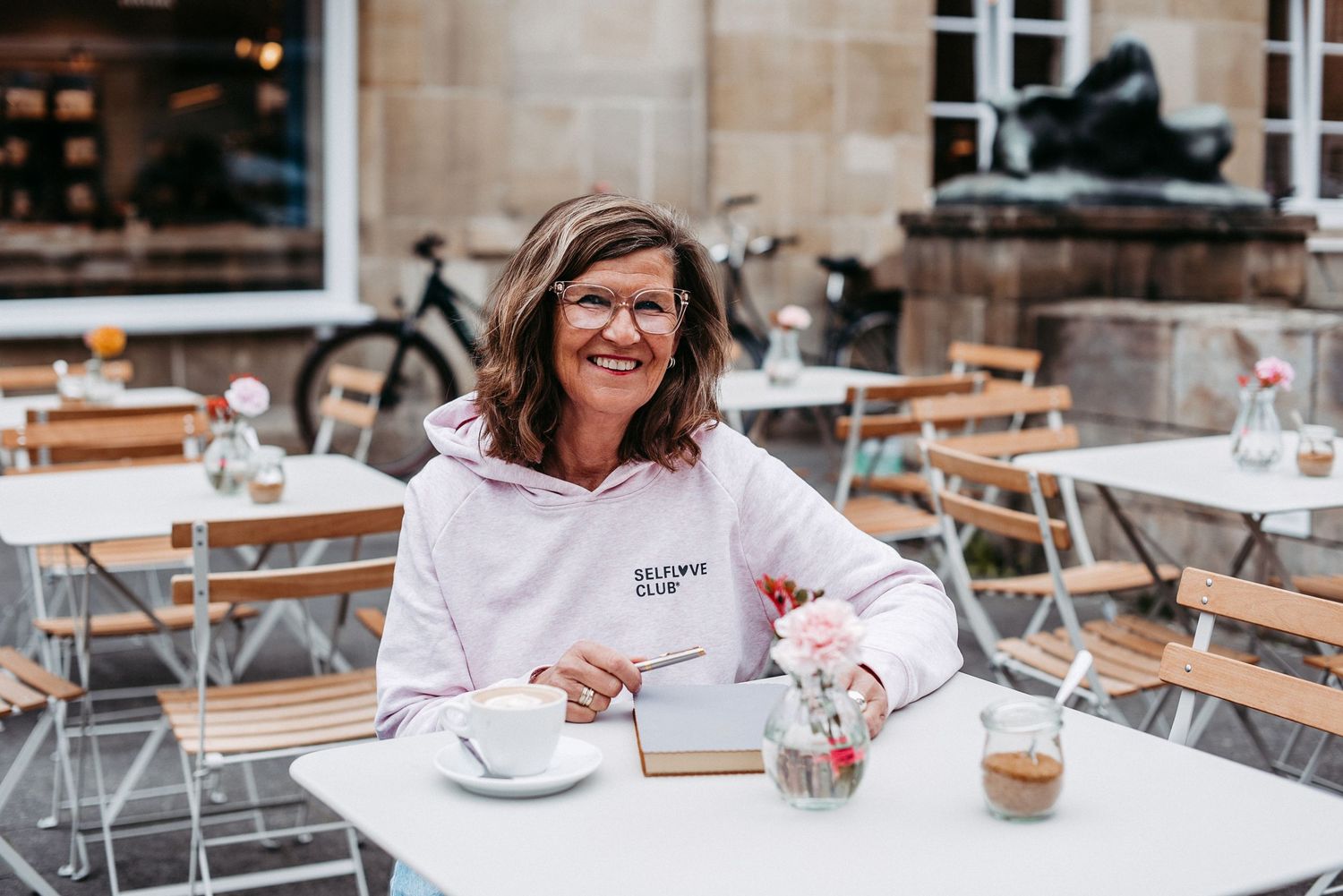 Frau mit Brille und rosa Hoodie sitzt lächelnd an einem Tisch im Freien. Im Hintergrund sind weitere Tische und ein Gebäude.