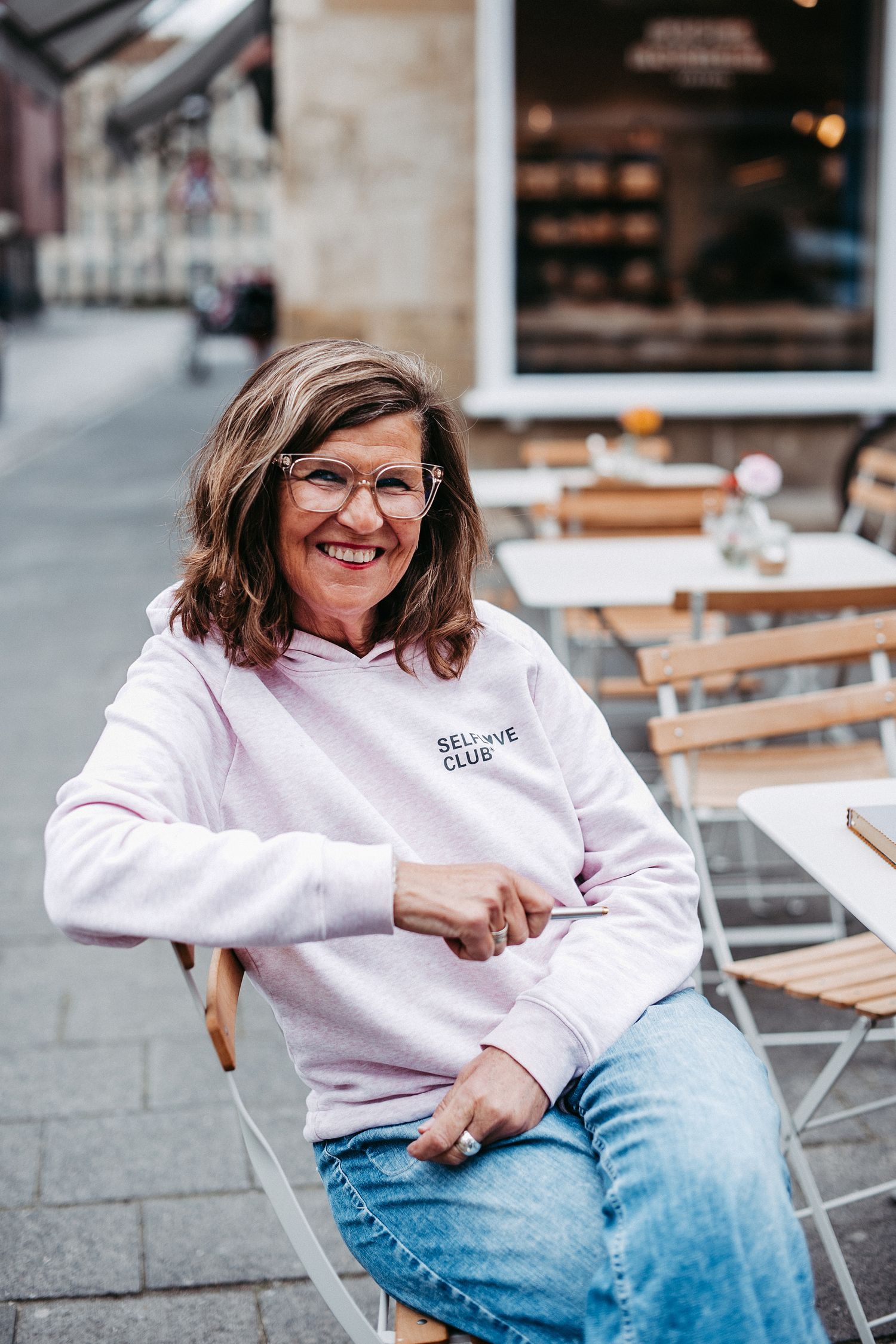 Frau mit Brille und rosa Hoodie sitzt lächelnd an einem Tisch im Freien. Im Hintergrund sind weitere Tische und ein Gebäude.