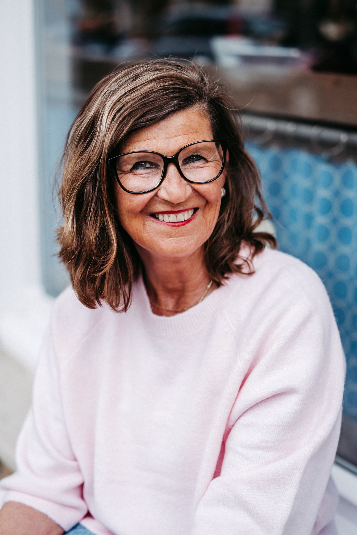 Frau mit Brille und rosa Hoodie sitzt lächelnd an einem Tisch im Freien. Im Hintergrund sind weitere Tische und ein Gebäude.