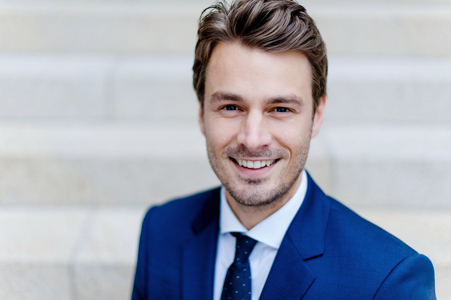 Ein lächelnder Mann in einem blauen Anzug steht vor einer Treppe. Er hat braune Haare und trägt ein weißes Hemd.
