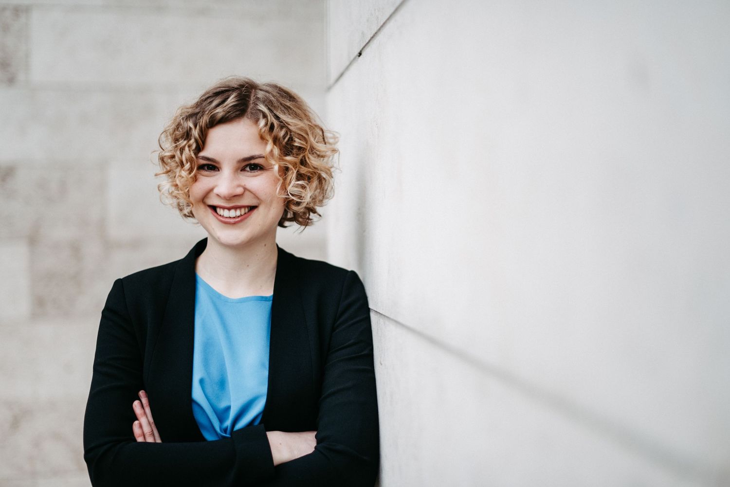 Businessfoto einer blonden jungen Frau mit lockigen Haaren, sie trägt eine blaue Blouse. Sie steht vor einer hellen Hauswand aus Beton.