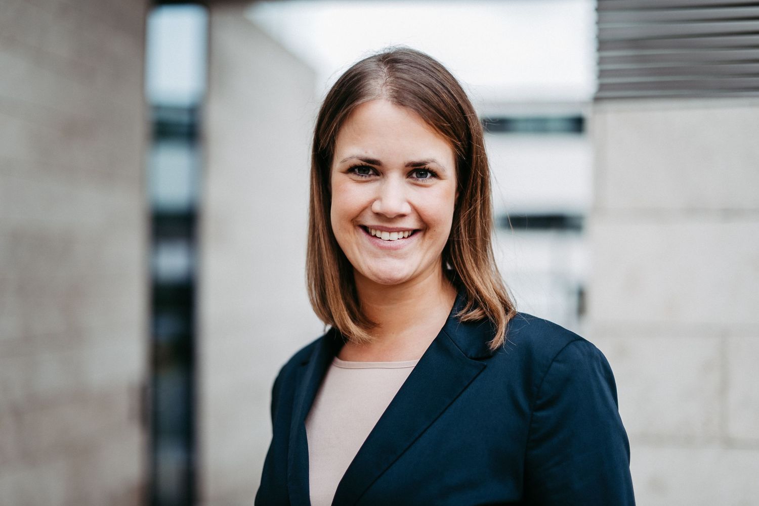 Eine lächelnde Person in einem dunklen Blazer steht vor einem modernen Gebäude mit Steinwänden und großen Fenstern.