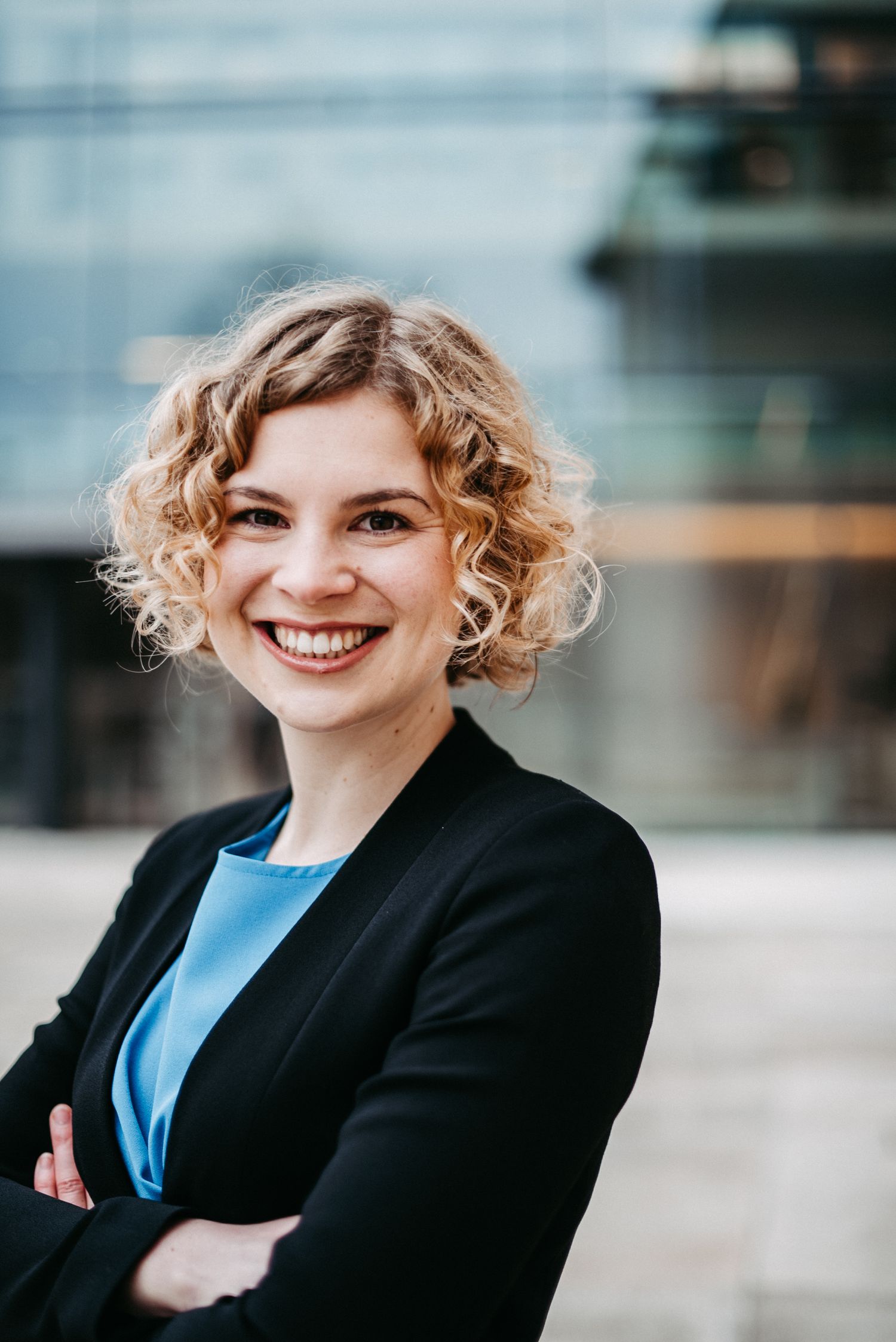 Lächelnde Frau mit lockigem Haar in einem schwarzen Blazer und blauer Bluse steht vor einem modernen Gebäude.