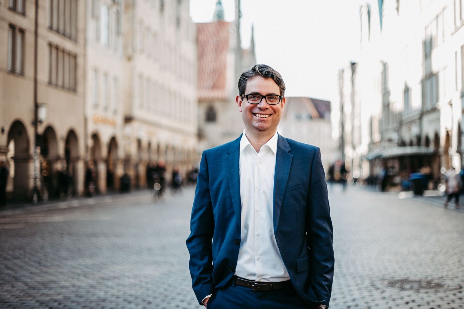 Businessfoto eines Mannes mit Blauem Anzug, weissem Hemd und Brille. Er lächelt in die Kamera. Im Hintergrund ist der Prinzipalmarkt.