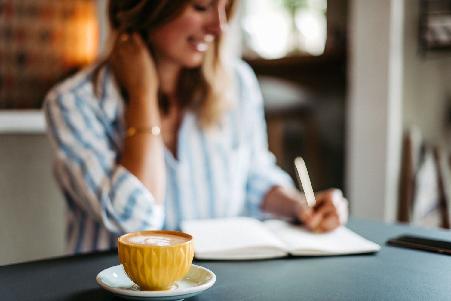 Eine Frau sitzt an einem Tisch und schreibt in ein Notizbuch. Im Vordergrund steht eine gelbe Tasse Kaffee auf einem Unterteller.