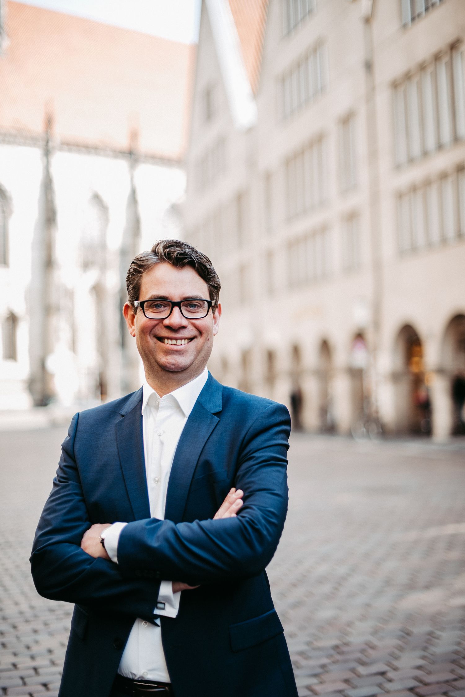 Eine lächelnde Person in einem blauen Anzug steht mit verschränkten Armen auf einem gepflasterten Platz vor historischen Gebäuden.