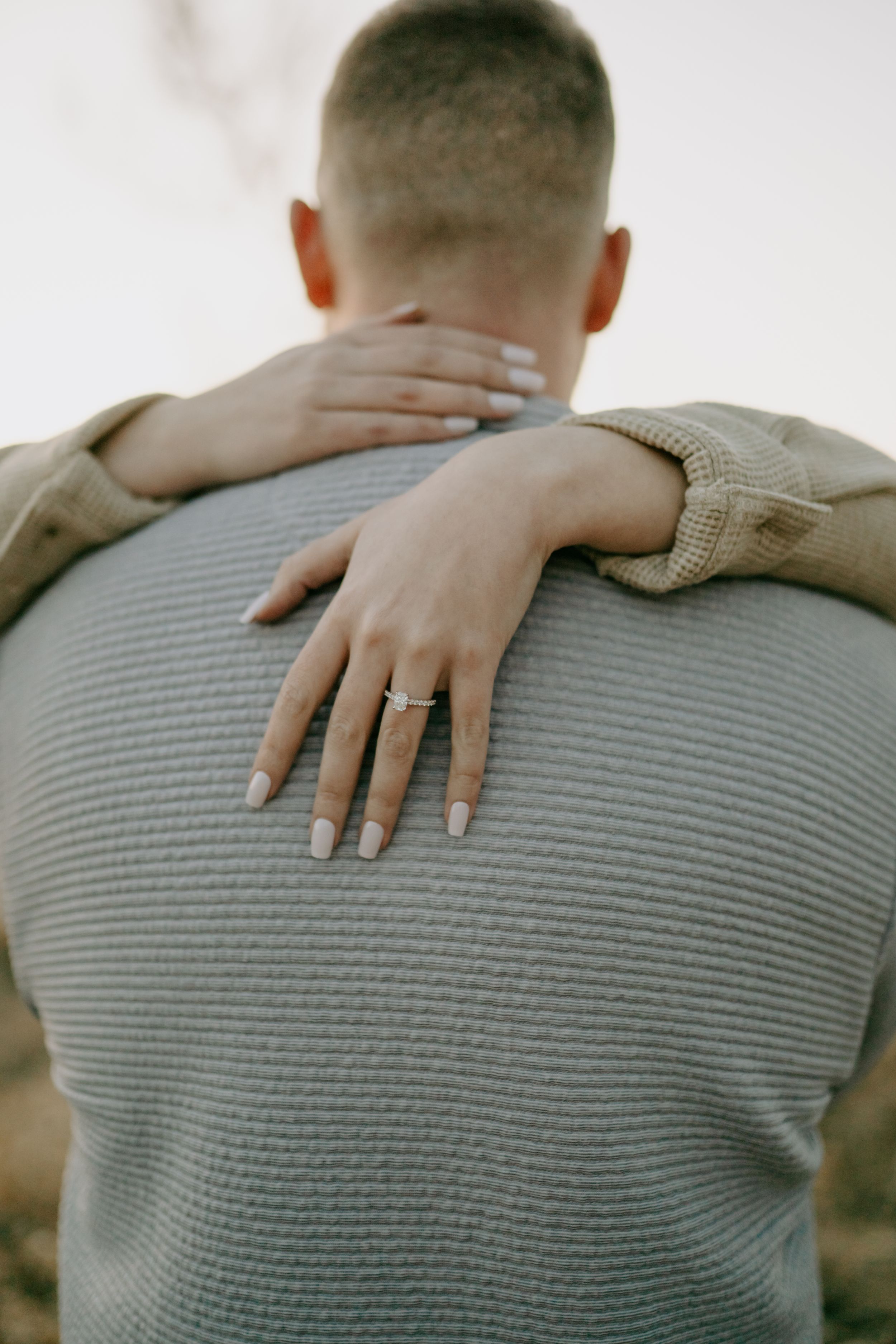 Close-up detail shot showing hands resting on a grey sweater with an engagement ring visible.