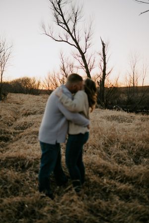 A couple embraces in a golden field with bare trees silhouetted against the sunset sky.