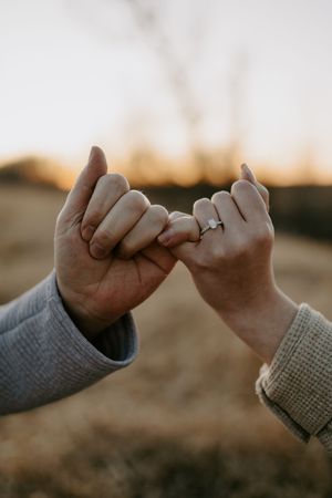 Two hands make a pinky promise against a warm sunset backdrop, with an engagement ring visible.