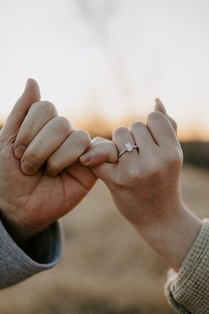 Close-up of intertwined pinky fingers with an engagement ring against a golden sunset.
