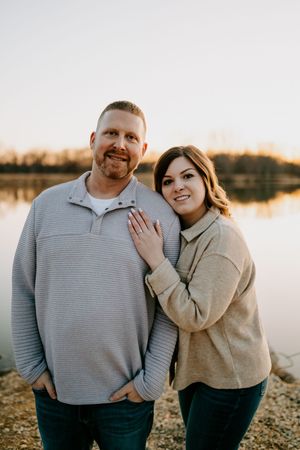 Happy couple in matching grey and beige sweaters posing by lake.
