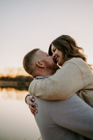 A romantic couple embraces during golden hour by a serene lake with warm sunset lighting in the background.