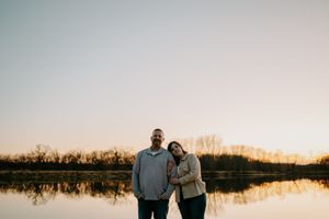 A couple stands together at sunset near a peaceful lake with trees in the distance.